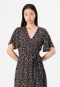 Robe noire à fleurs avec un décolleté en V profond, des manches courtes en chandelier et une taille à nouer. Elle présente un tissu texturé avec de petites fleurs roses et blanches.