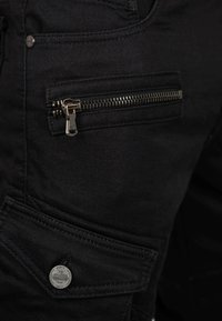 Primo piano di pantaloni in tessuto nero con una tasca orizzontale con cerniera metallica e una tasca con patta abbottonata con incisione del marchio.