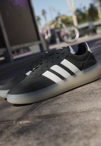 Sneakers Adidas bianche e nere con suole traslucide posizionate all'aperto su un pavimento in pietra vicino a una bicicletta.
