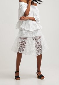 Bílá blůza s odhalenými rameny a peplum designem, zkombinovaná s vrstvenou, průhlednou bílou krajkovou sukní s složitými vzory. Černé lodičky s kotníkovými pásky.