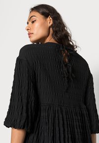 Vestido preto com pregas e tecido texturizado, mangas bufantes e fecho de laço nas costas. Apresenta um corte solto e uma bainha com babados para um detalhe adicional.