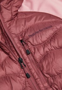 Zbliżenie na pikowaną tkaninę kurtki w kolorze zakurzonego różu z dwoma suwakami i wyhaftowanym logo "Peak Performance" na piersi.