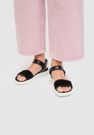 Füße, die schwarze Sandalen mit offenen Zehen, weißen Sohlen und Knöchelriemen tragen, kombiniert mit weit geschnittenen rosa Hosen, die oberhalb der Knöchel enden.