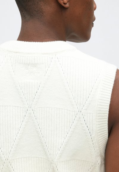 Suéter sin mangas de punto blanco con un patrón texturizado de diamantes y un cuello acanalado, que muestra un acabado suave y una tela blanda.