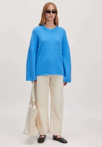 Heldere blauwe, oversized gebreide trui met een ronde hals, gecombineerd met crèmekleurige wijde broek en zwarte platte schoenen. Inclusief een beige draagtas.
