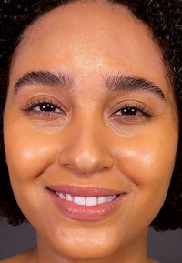 Nærbilde av en persons ansikt med glatt, strålende hud, mørkt krøllete hår, naturlig sminke og et subtilt smil. Har klare øyne og definerte bryn.