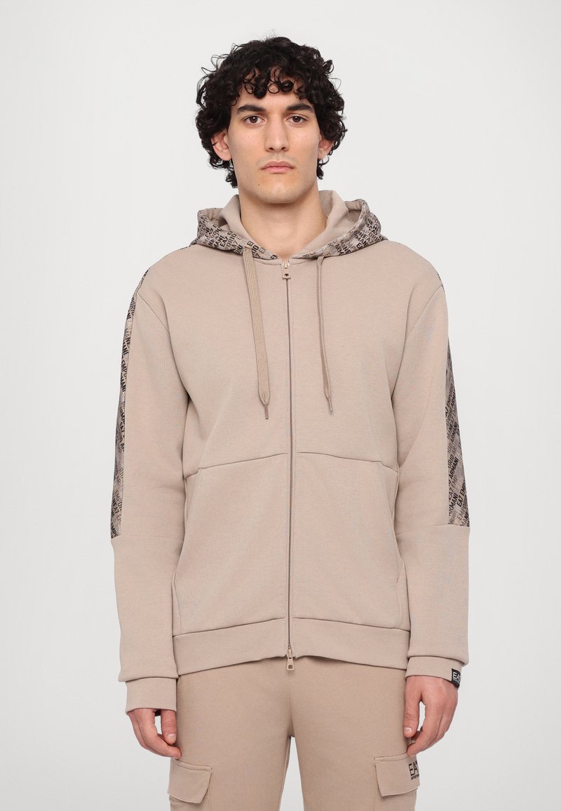 Sudadera con cremallera en color beige, hecha de un tejido suave, con capucha estampada y paneles laterales, además de bolsillos tipo canguro y cordones ajustables.