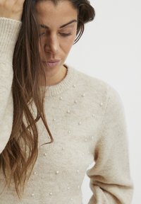 Vaaleanbeige neulepaita pyöreällä kaula-aukolla, jossa on hajallaan valkoisia helmiä ja ribbireunukset. Pehmeä tekstuuri, rento istuvuus.