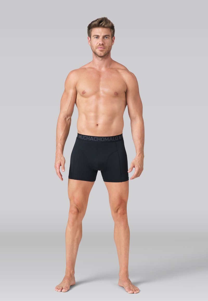 Gesfitte man staat blootsvoets en draagt zwarte boxershorts met een merkbare tailleband tegen een effen grijze achtergrond.