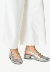 Strieborné metalické slingback topánky so štvorcovou špičkou, vybavené dvoma nastaviteľnými popruhmi a nízkym blokovým podpätkom. Nosené s bielymi širokými nohavicami.