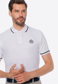 Bílá polo tričko s černým límcem a akcenty na rukávech, obsahující tři knoflíky a vyšívané logo na hrudi. Měkká, texturovaná tkanina.