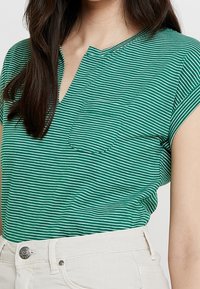 Vihreä raidallinen lyhythihainen paita V-kaula-aukolla ja taskulla. Kangas on sileää ja siinä on vaakasuoria raitoja eri leveys.