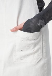 Biały dżinsowy fartuch z koronkowym detalem w kieszeni; kieszeń ma małe wycięcia i czerwone akcenty. Gładka tekstura, casualowy design.