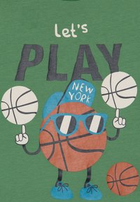 Camiseta verde con un personaje de baloncesto de dibujos animados, que lleva gafas de sol y una gorra etiquetada "NEW YORK", con el texto "vamos a JUGAR" y balones de baloncesto.