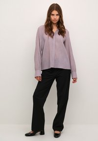 Blusa lunga a maniche con righe verticali di colore viola chiaro e scollatura a V, abbinata a pantaloni neri a gamba larga e scarpe nere con tacco.