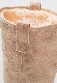 Beżowy kozak z ekoskóry z miękkim, beżowym futerkiem w środku, z prostym pętelkowym zdobieniem na cholewce i gładką, o fakturze zewnętrzną.