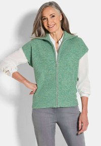 Gilet en tricot côtelé vert avec un col montant et une fermeture éclair. Porté sur une chemise blanche, associé à un pantalon gris clair. Texture lisse et coupe décontractée.