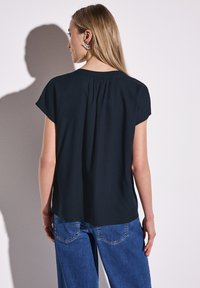 Blusa blu scuro a maniche corte, con dettaglio a pieghe sul retro, abbinata a jeans a gamba larga blu chiaro. Tessuto liscio con una vestibilità comoda.