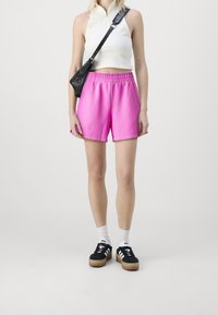 Haut court blanc ajusté avec une fermeture éclair, associé à un short ample rose vif. Sac bandoulière noir et baskets noires avec des accents blancs.