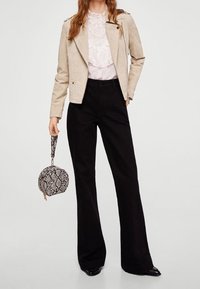 Donna che indossa un giubbotto beige, una camicetta di pizzo bianca, pantaloni a gamba larga neri, mentre tiene una borsa rotonda con stampa serpente per la maniglia.