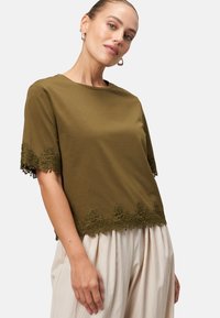 Olive green short-sleeve top in soft cotton, featuring a scalloped lace trim at the hem. Paired with light, pleated trousers.