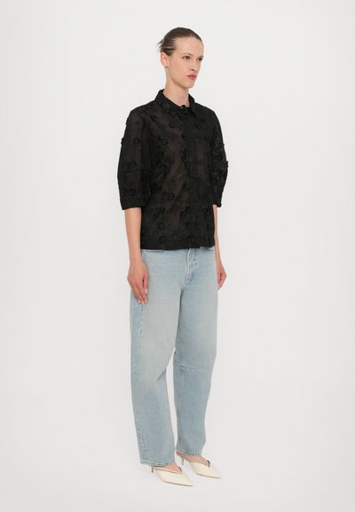 Sort gennemsigtig bluse med blomsterbroderi, knaplukning foran og ærmer, der går til trekvartlange. Den er parret med lyseblå rummelige jeans og spidse hvide hæle.