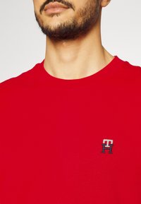 T-shirt di cotone rosso con scollatura tonda e un piccolo logo ricamato in colori a contrasto sul petto sinistro. Tessuto morbido.