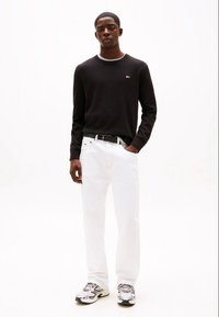 Black long-sleeve sweater with a round neckline, white jeans, and grey sneakers featuring black and white accents, standing against a light background.