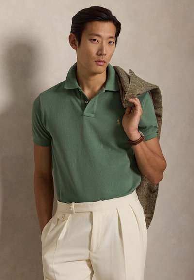 Vyras, vilkintis žalią polo marškinėlį ir kreminius plyšiuotus kelnes, laiko raštuotą švarką ant peties paprastame smėlio spalvos fone.