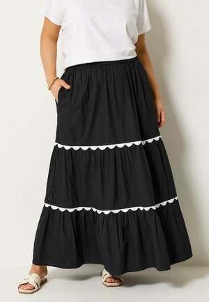 Kvinde iført en sort lagdelt maxinederdel med hvid scallopkant, hvid t-shirt, guldbøjle og hvide slip-on sandaler.