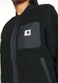 Mustat fleece-takki, jossa on teksturoitu pinta, sileä nailonrintatasku, vetoketjukiinnitys ja brodeerattu logo.