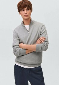 Ljust grå zip-up tröja med ribbad krage och ärmmuddar, lagrad över en vit T-shirt, bärs med mörkblå byxor. Slät tygtextur.