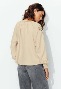 Blouse beige à manches longues avec broderie florale sur les épaules, poignets élastiques, tissu texturé et coupe décontractée. Assortie à un jean gris.