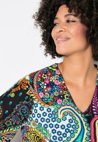 Vestido colorido y con estampados con escote en V, que presenta una mezcla de diseños florales en azul, rosa y verde sobre una base negra. Cabello rizado visible.