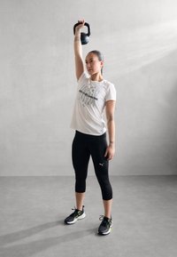 Leggings de sport noirs, t-shirt blanc avec du texte, kettlebell gris levé au-dessus de la tête. Baskets noires et vert lime avec des lacets. Fond en béton.