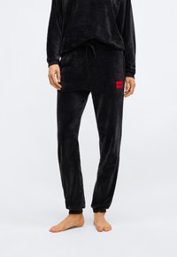 Zwart velours lounge-outfit bestaande uit een losse, langarmoenen top en taps toelopende broek met een verstelbare koordtaille en een rood logo-patch.