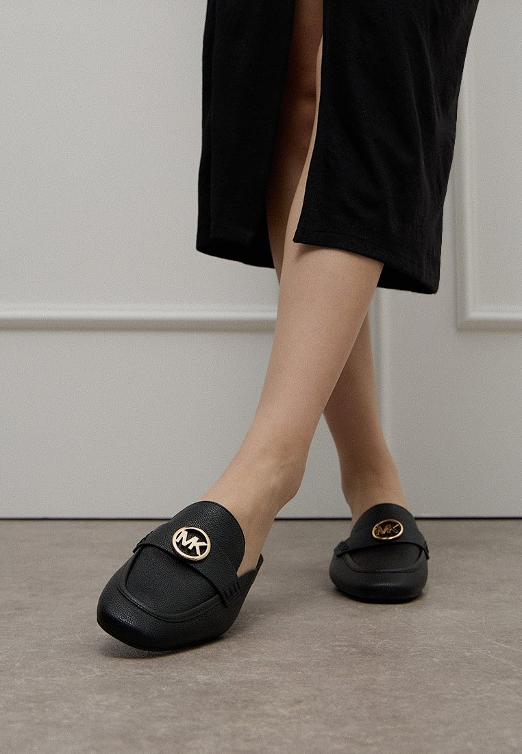 Zwarte leren instappers met een ronde neus, voorzien van een opvallend gouden logo aan de voorkant en subtiele stiksels aan de zijkanten.