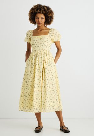 Frau steht mit den Händen in den Taschen, trägt ein gelbes Midikleid mit Blumenmuster, Puffärmeln und quadratischem Ausschnitt, kombiniert mit schwarzen Ballerinas.