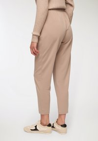 Beżowe spodnie o luźnym kroju z elastycznym paskiem w talii, zwężane u kostek i o miękkiej teksturze, zestawione z jasnymi sneakersami z czarnymi akcentami.