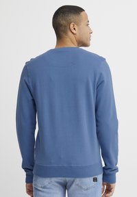 Sudadera azul con mangas largas y un escote redondo. Textura suave y un dobladillo acanalado. Detalle de etiqueta lateral visible cerca de la cintura.