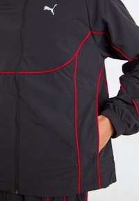 Svart treningsjakke med røde detaljer, med glidelås foran, elastiske mansjetter, og en liten hvit logo på brystet. Lett materiale.