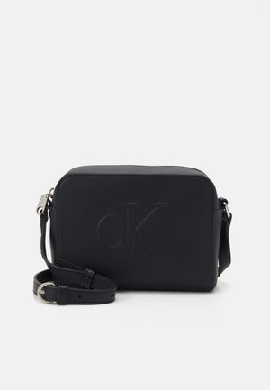 Sort rektangulær crossbody taske med justerbar strop og præget "cK" logo på forsiden, sat mod en ensfarvet lys baggrund.