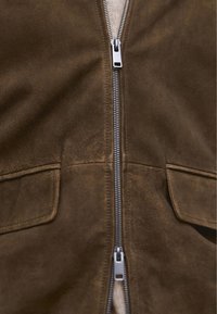 Brun ruskindjakke med sølv lynlås, to frontlommer og en glat overflade. Indvendigt lag i en lysere farve.