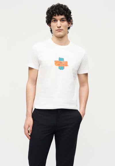 Άνδρας με σγουρά μαλλιά που φοράει λευκό μπλουζάκι με γαλάζιο και πορτοκαλί αφηρημένο σχέδιο και μαύρο παντελόνι, στέκεται με το ένα χέρι στην τσέπη.