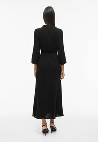 Juodas ilgomis rankovėmis maksi suknelė su prigludusia juosmens dalimi, pasižyminti lygiu paviršiumi ir vėjinga siluetu. Juodi aukšti kulniukai su dirželio detalėmis.