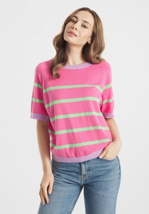 Jonge vrouw met golvend haar, gekleed in een roze trui met groene strepen en lichtpaarse rand, gecombineerd met een blauwe spijkerbroek, poserend met één hand in haar zak.