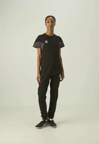 Fekete sportos póló szürke részekkel és logóval a mellkasnál, fekete nadrággal és fekete sportos cipővel párosítva. Egyszínű háttér.