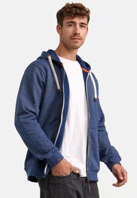 Sudadera con capucha de color azul oscuro, con una superficie texturizada, cremallera completa, cordones crema y bolsillos laterales. Llevada sobre una camiseta blanca.