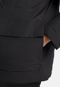 Veste noire en tissu lisse et texturé, avec une poche sur le devant et une coupe ample. Le design met l'accent sur la praticité et le confort.