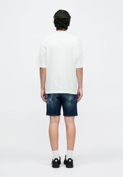 Camiseta blanca de manga corta combinada con pantalones cortos de mezclilla azul. Los pantalones cortos tienen detalles desgastados y bolsillos. La modelo lleva zapatillas negras y calcetines blancos.